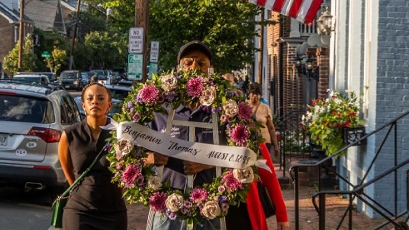 Procession with wreath passing house with flag.