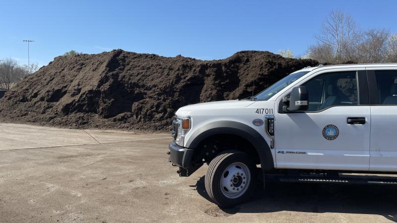 A white pickup truck with the city seal on it parked in front of a large pile of dark brown mulch.