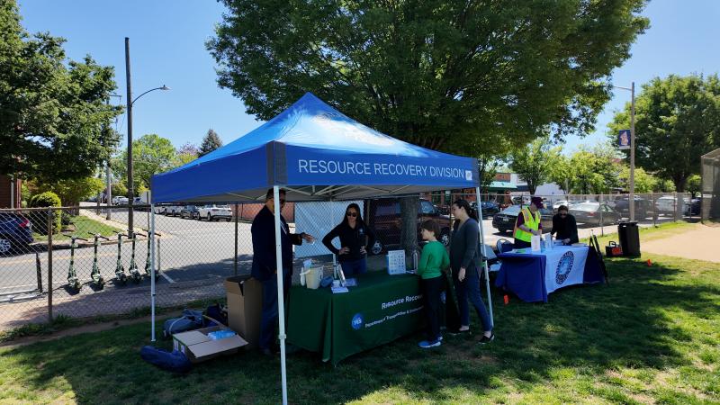 A photo of a pop up tent with "Resource Recovery" written on the top, with a table and two city staff members underneath. They are speaking with residents.