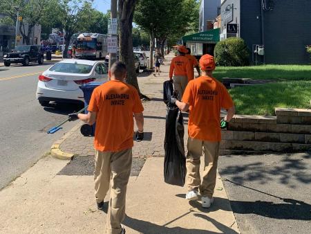 four people visible from behind wearing bright orange shirts that say Alexandria Inmate and collecting litter from a sidewalk along Mount Vernon Avenue