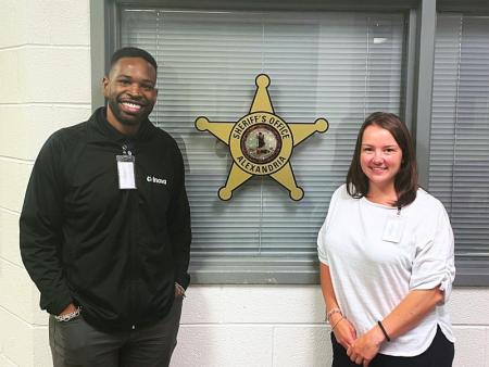 Two public health professionals, smiling, in front of a large star-shaped window decal that says Sheriff's Office Alexandria