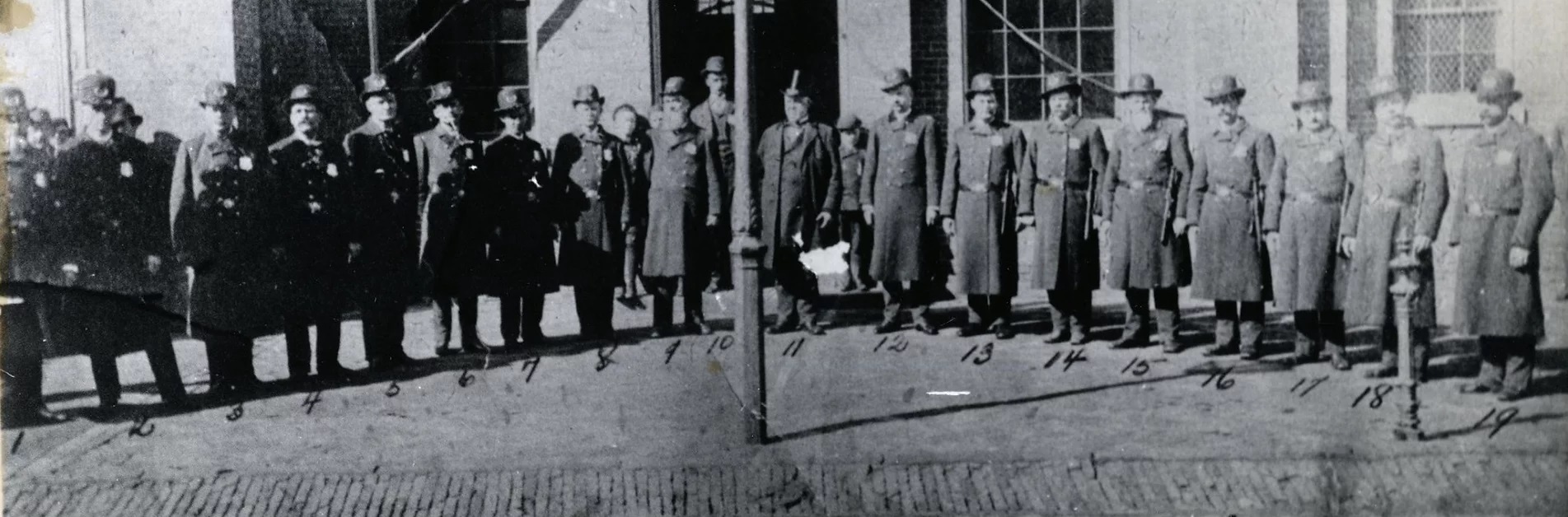 A black and white photo of men in police uniforms standing in a single line in front of a brick building
