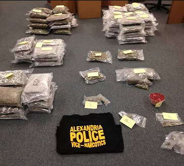 A folded t-shirt on a carpeted floor that reads "Alexandria Police Vice Narcotics". Surrounding the t-shirt are bags of what appear to be seized drugs.