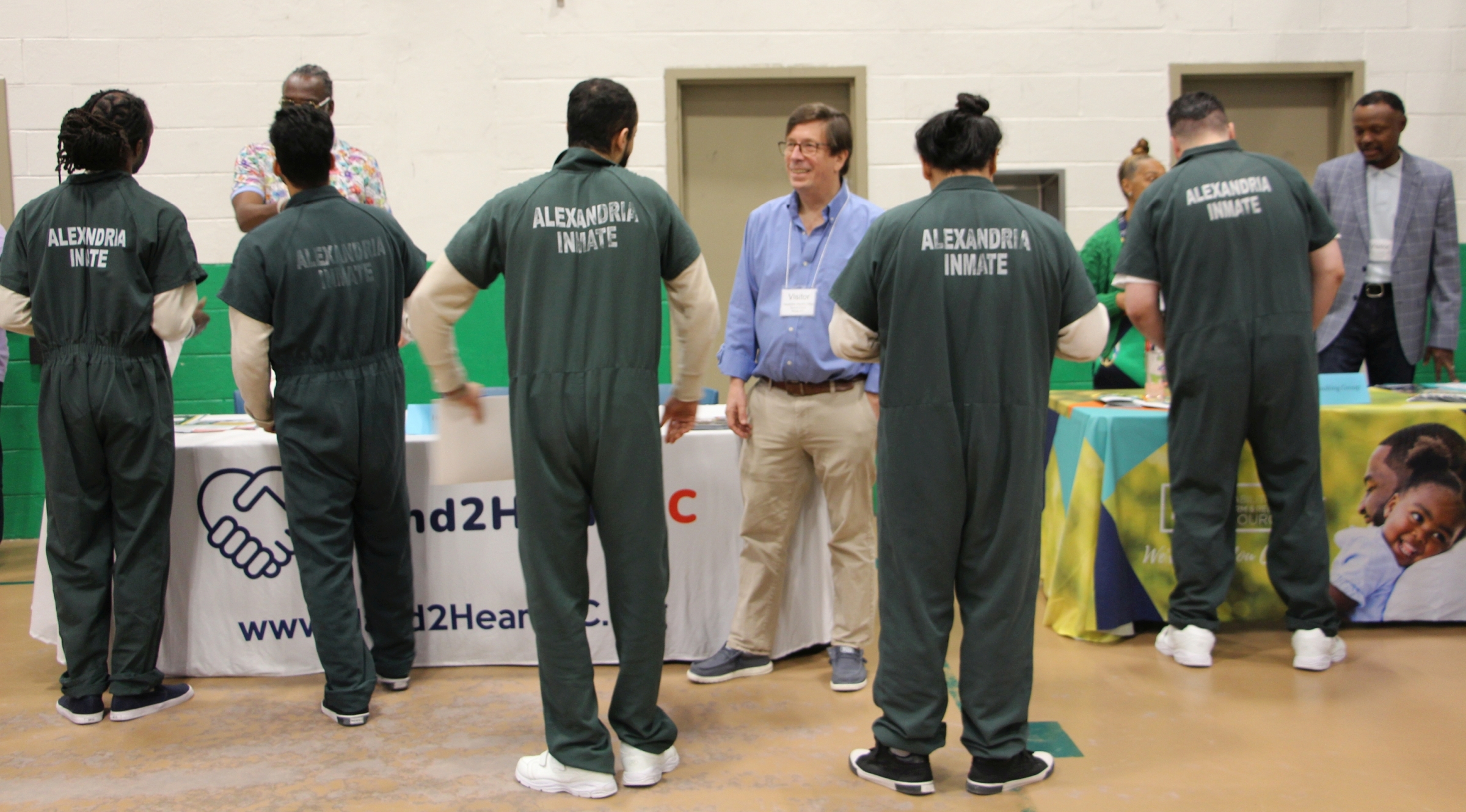 Five inmates in green jumpsuits speaking with four individuals from two diferent organizations and looking at information at exhibition tables
