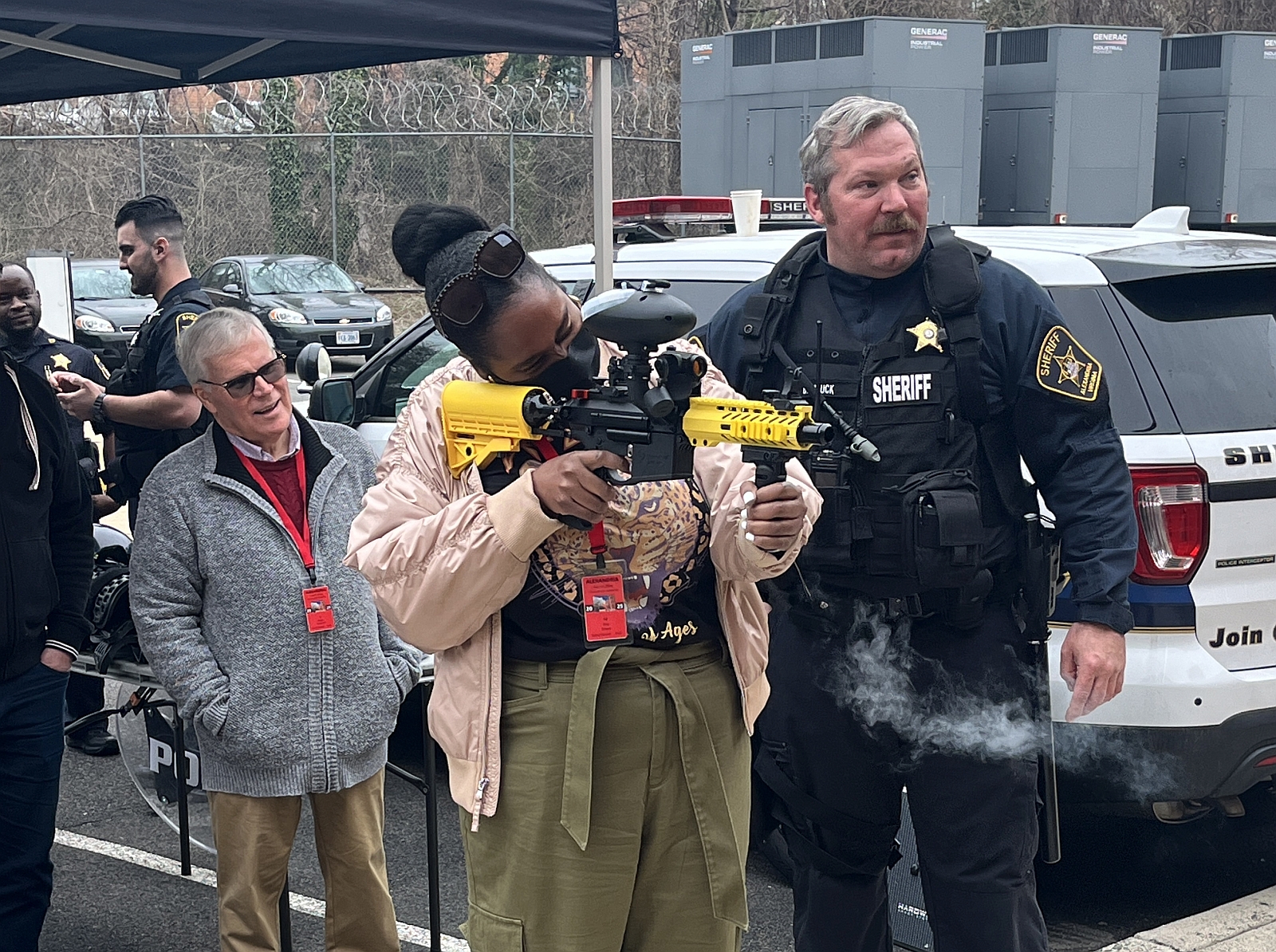Civilian firing a pepperball training launcher under the supervision of a Sheriff's sergeant in a blue uniform.