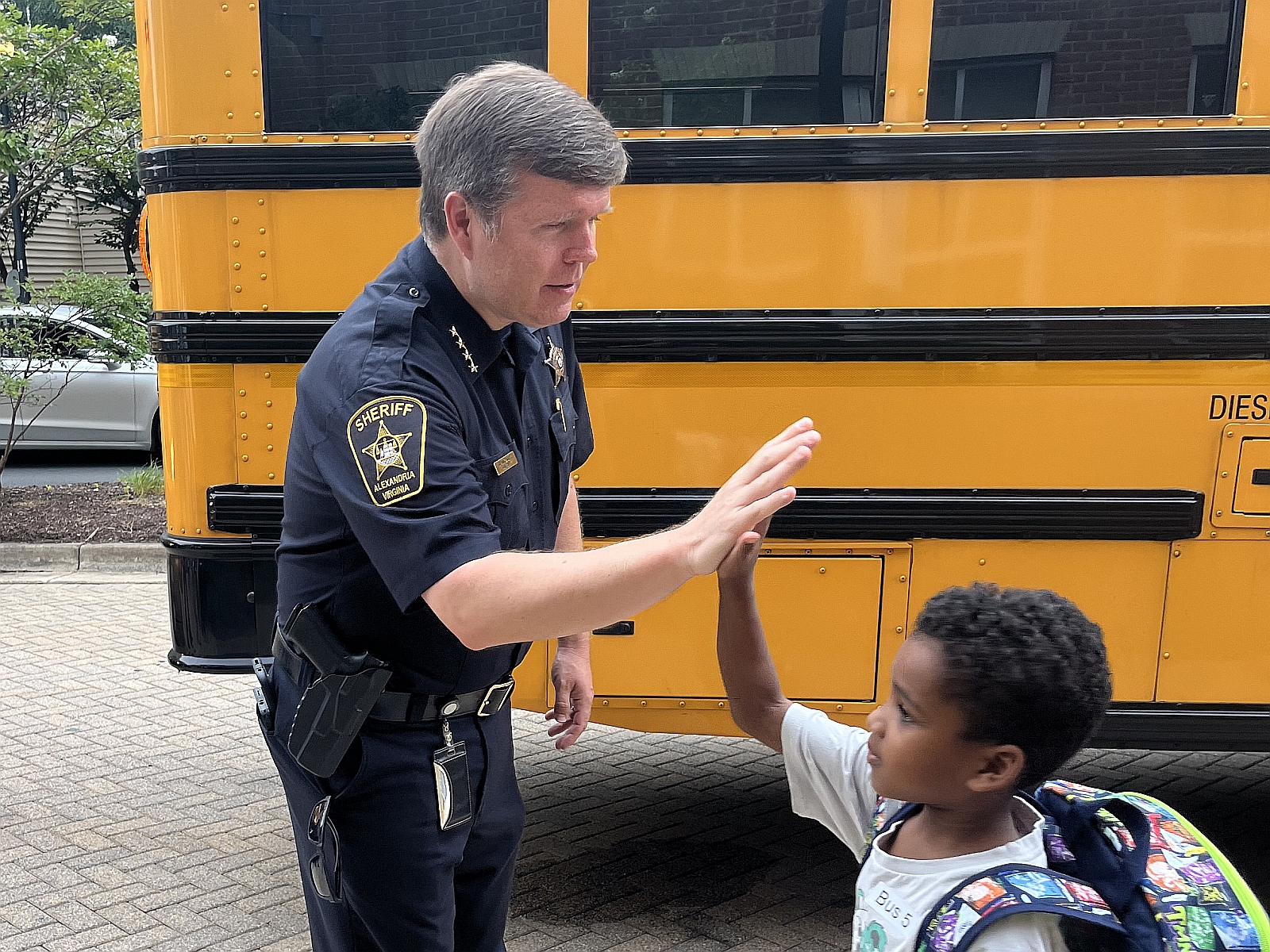 Sheriff in a blue uniform high fiving an elementary school student on the first day of school with a school bus in the background