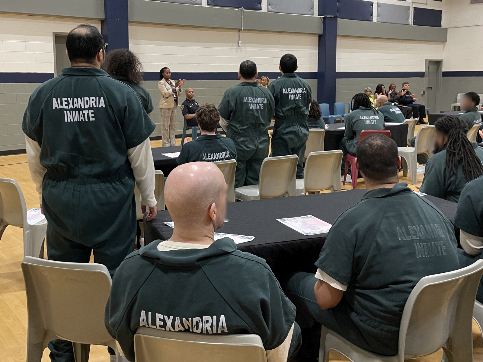 Several inmates in green jumpsuits, some standing and seated, in a gymnasium visible from behind