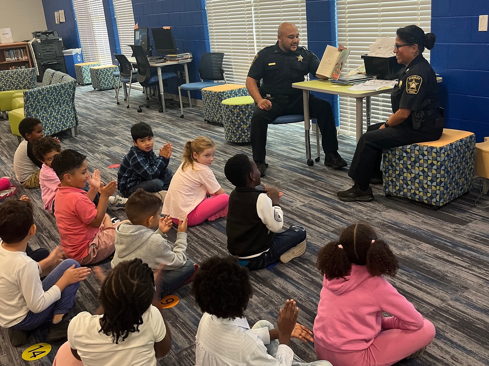 Two deputies in blue uniforms reading books to elementary school students who are sitting on the floor