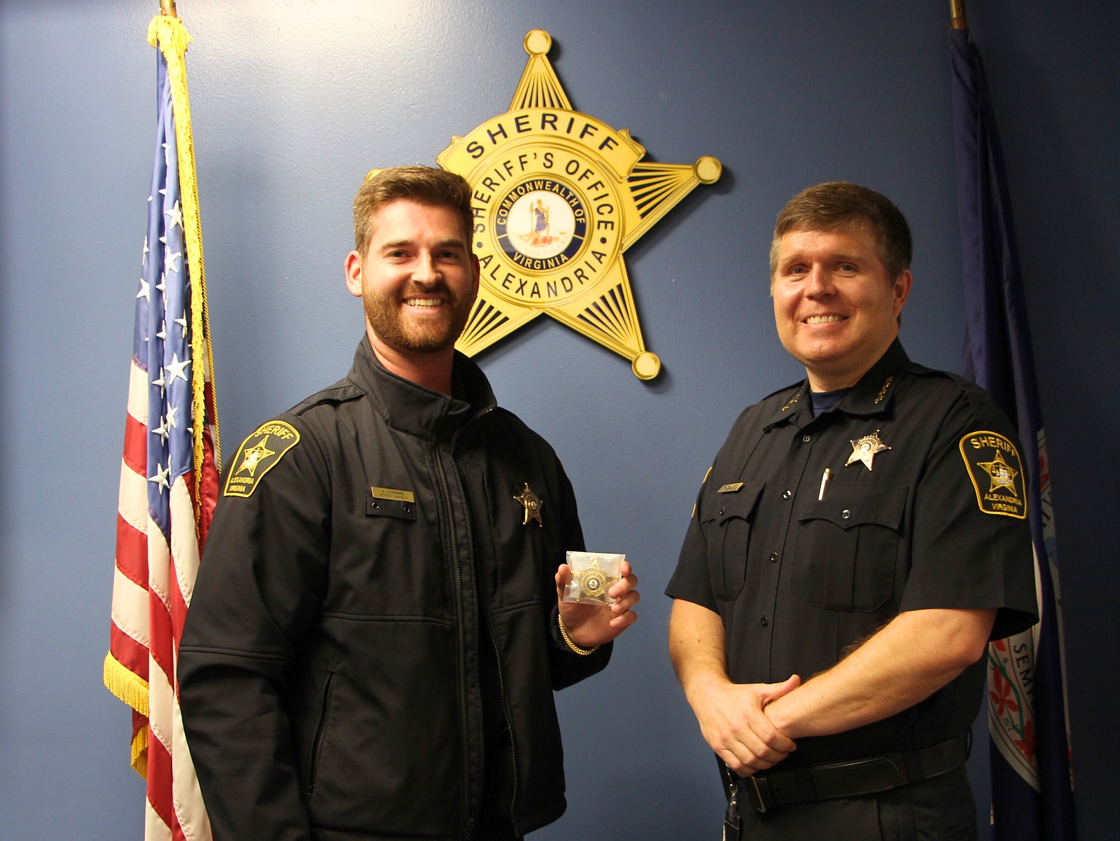Deputy and Sheriff, both in blue uniforms, standing in front of a U.S. and a Virginia flag with a gold sheriff's star on the wall