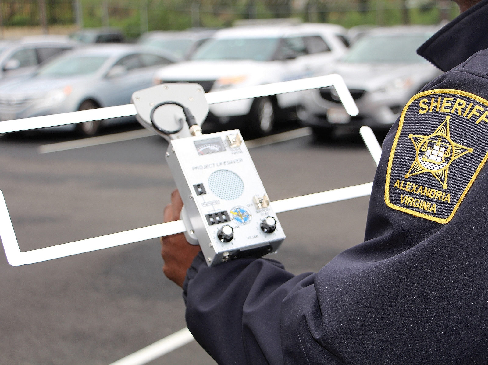 A silver colored metal tracking device being held by a deputy whose blue jacket sleeve and should patch are visible