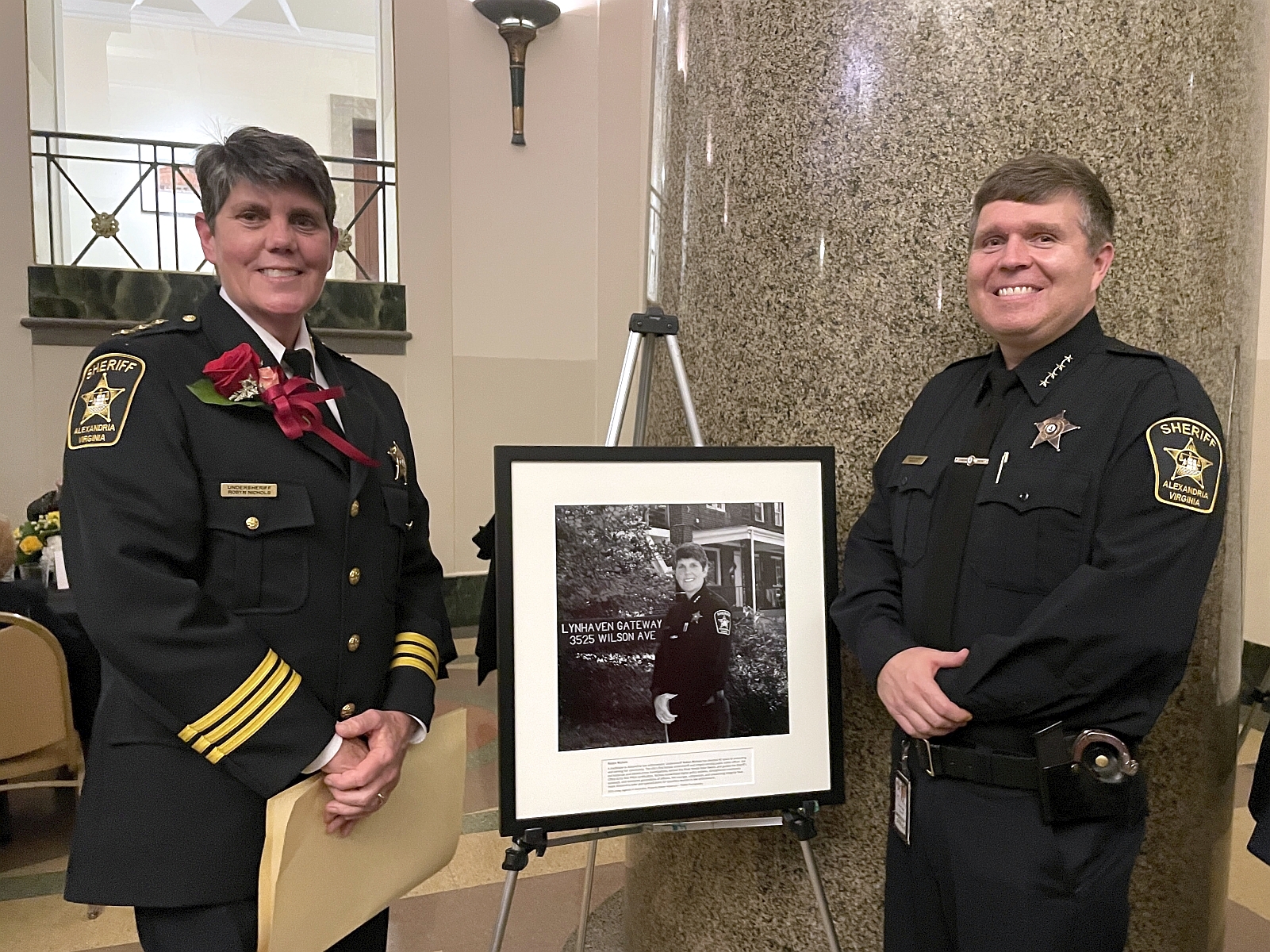 Undersheriff on left and Sheriff on right, both in blue uniforms, standing with a framed black and white photograph of the undersheriff between them