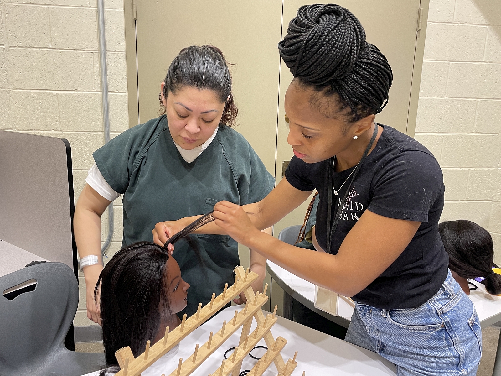 Inmate in green uniform looking closely as a braiding instructor demonstrates a braiding technique on a mannequin head