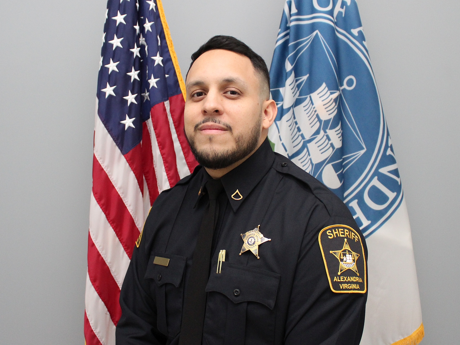 Official portrait of Deputy Gonzalez wearing blue uniform with U.S. and Alexandria flags in background