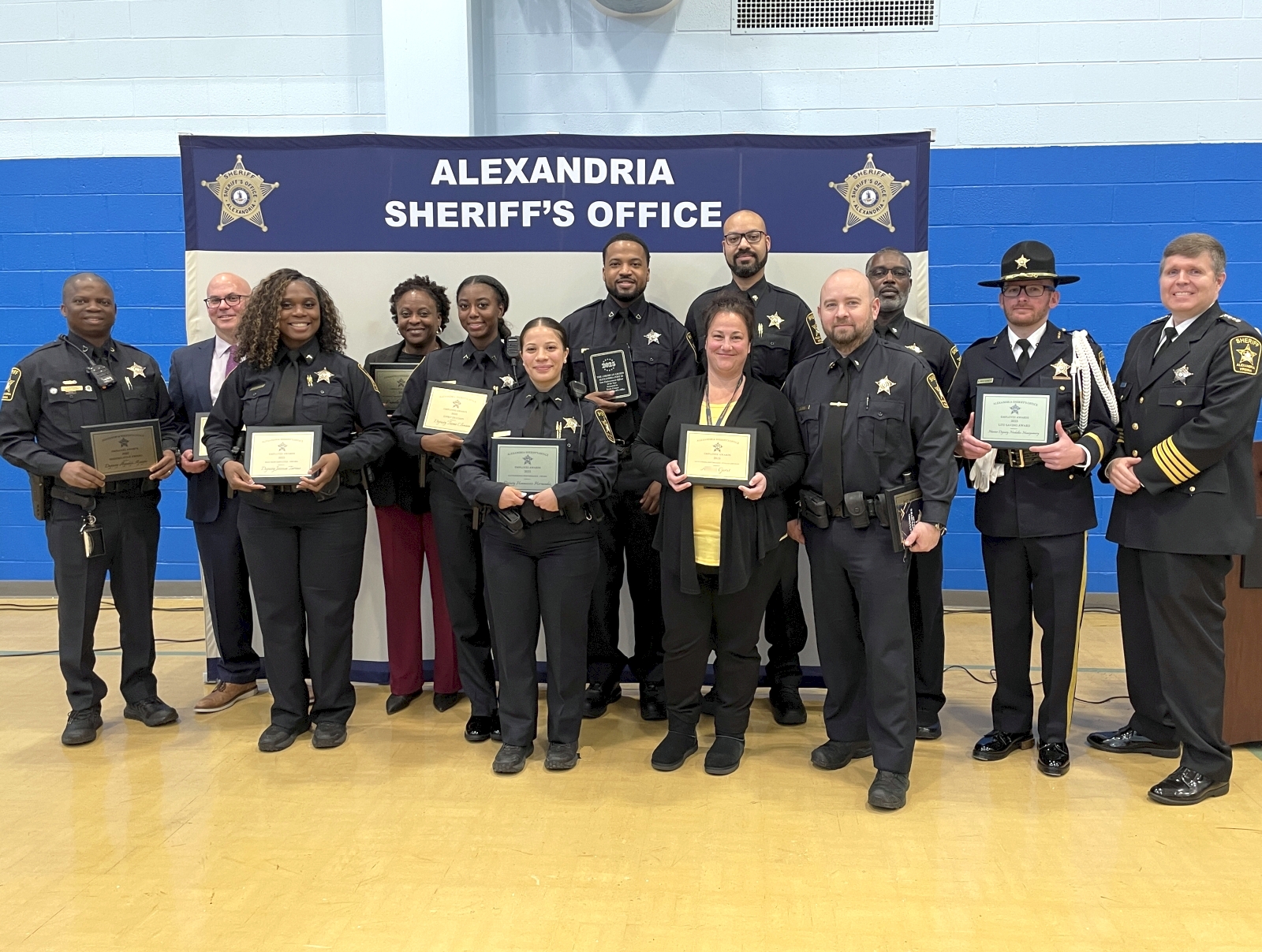 Deputies and civilian staff holding plaques and smiling at an awards ceremony in front a backdrop that says Alexandria Sheriff's Office