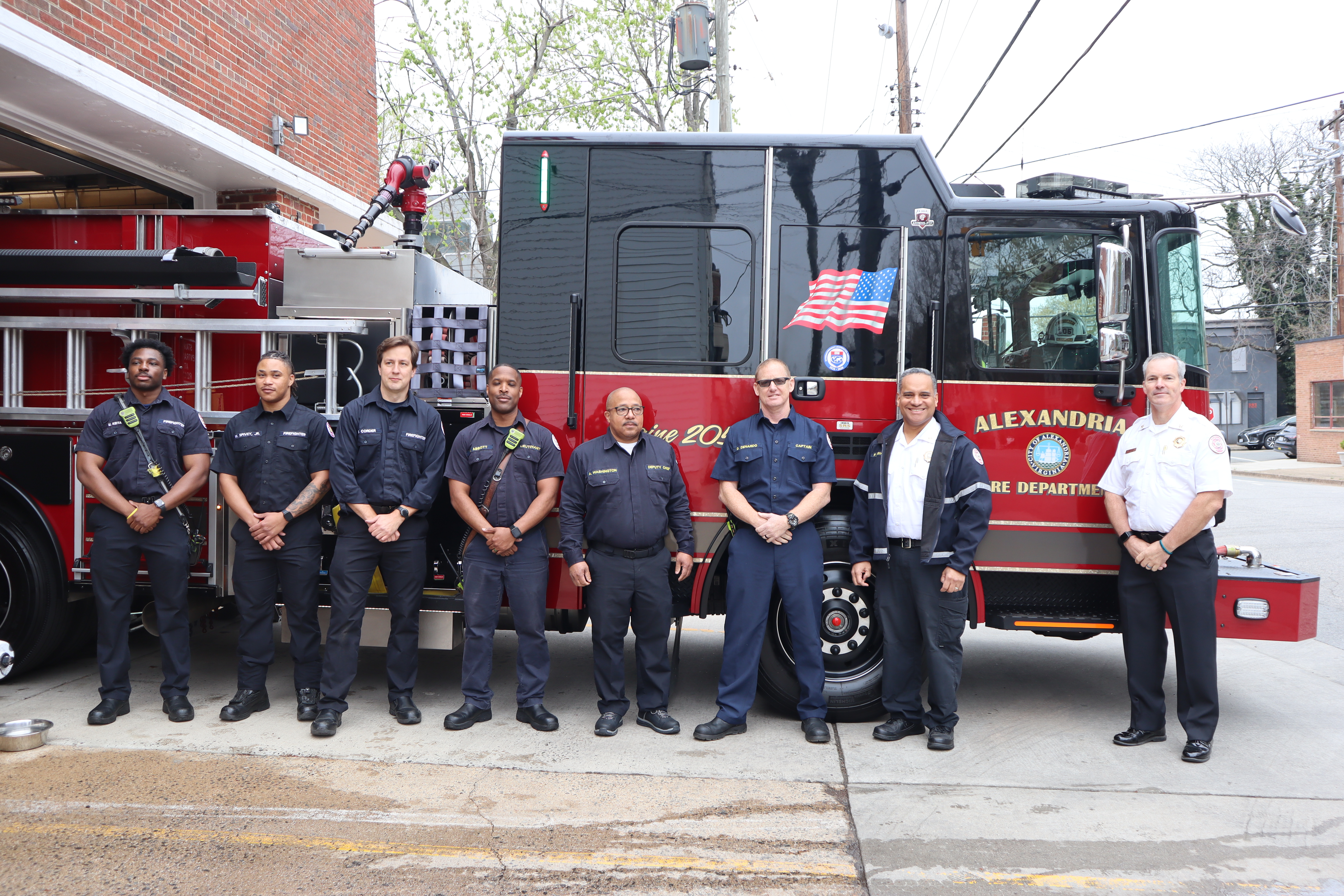 A group of people in fire department uniforms stand in front of an Alexandria Fire Department truck. There are eight individuals, all posing and smiling for the photo in an outdoor setting.