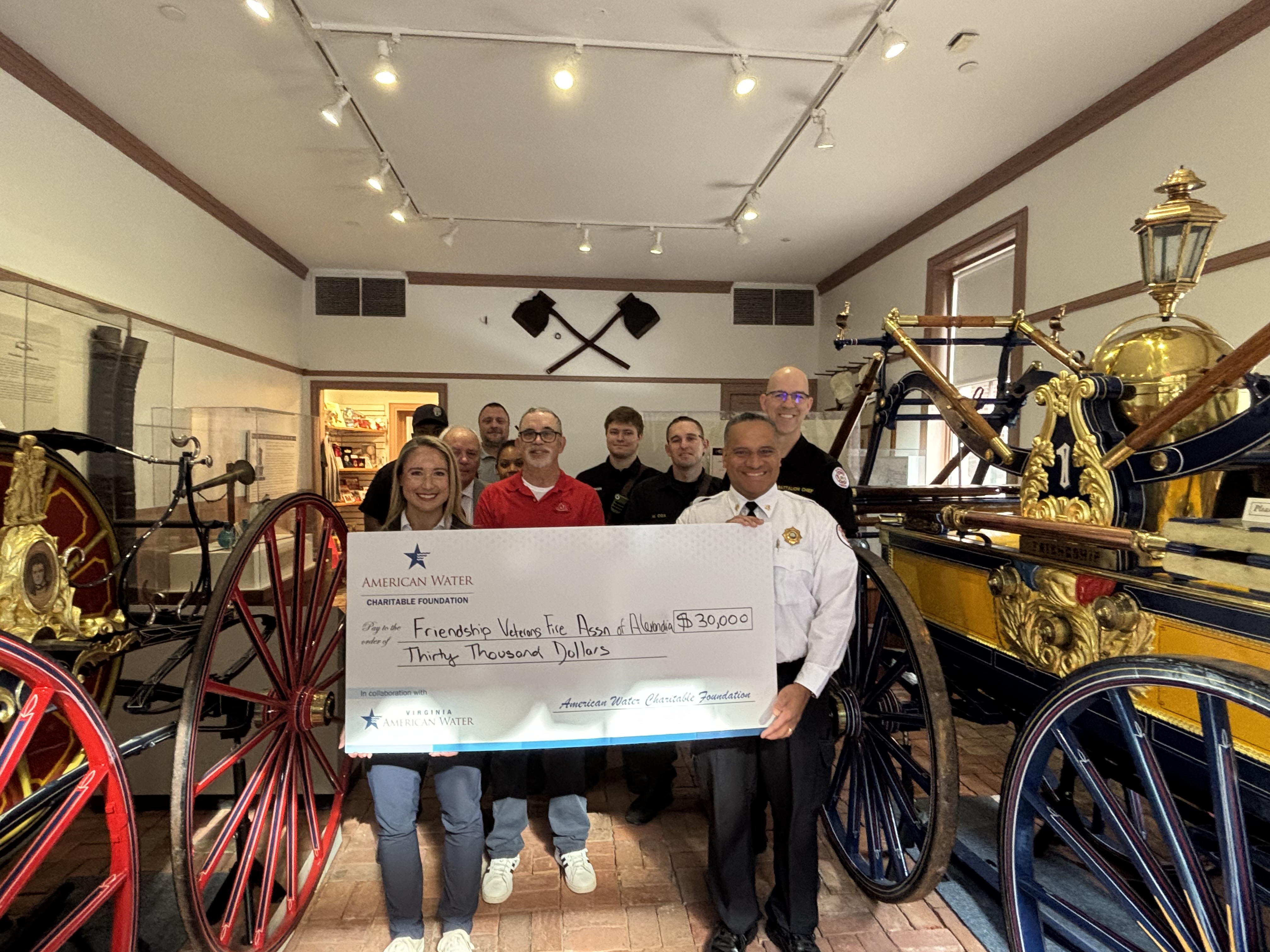 Group of 10 people smiling holding a large check from American Water. The amount is for thirty thousand dollars. The group is standing between two vintage fire vehicles. They are in a room with lights hanging above and two axes displayed on the wall behind them.