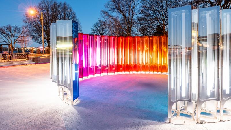 Mirror Mirror's lights shine at dusk Outside the nearly enclosed circle are white, mirrored lights. Inside the red, pink, and orange lights of the rainbow interior can be seen.