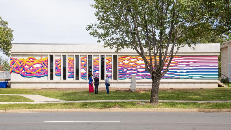 Confluent Threads mural as seen from across Seminary Road. Bands of colored lines converge in an intertwining pattern as the mural moves from the back right corner to the left and toward the entrance of the library,