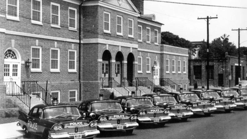 A black and white photo of a row of dark cars with POLICE written on the front of them in white letters, parked outside of a brick building.