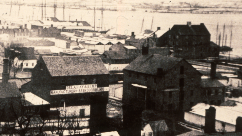 A black and white photo shows buildings along the waterfront with the masts of tall ships in the background. The Jamieson Bakery building is a three-story building with loft, circled in the foreground.