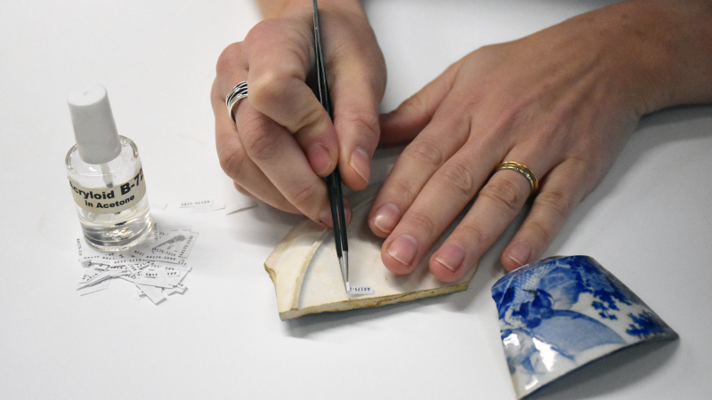 An archaeologist uses tweezers and a removeable glue to add small paper labels to the base of a ceramic artifact