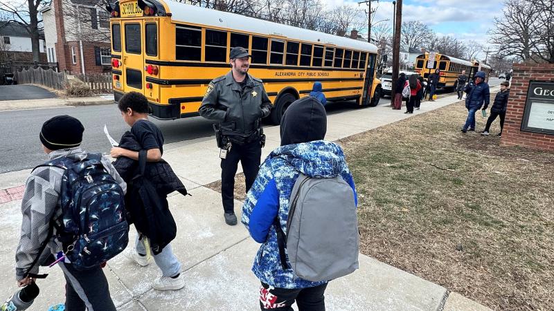 Deputy in a blue uniform watching as elementary students during dismissal. An orange school bus is in the background.