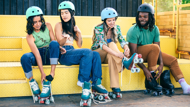 Four teens with helmets on sitting on yellow bleachers, while putting on rollerblades