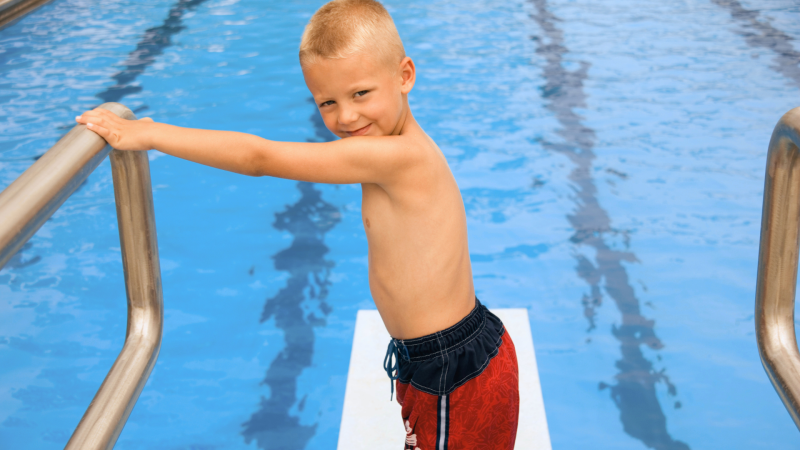 Blonde boy standing on a diving board while holding onto a rail