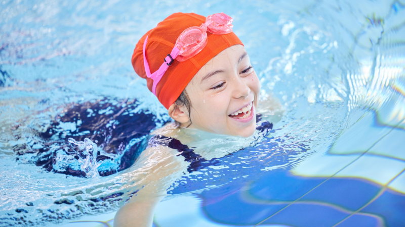 Smiling female child with red swim cap and pink goggles swimming in an indoor pool