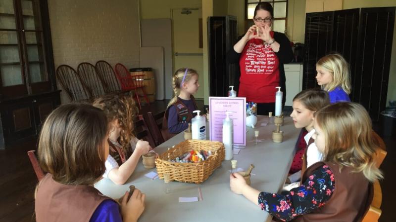 Brownie Girl Scouts are seated around a table listening to craft instructions from museum staff wearing a red shirt. 