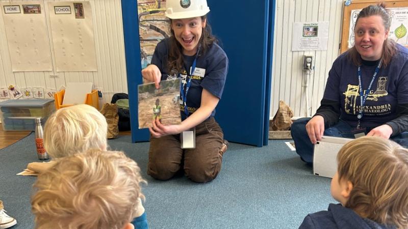 A museum educator in a blue Alexandria Archaeology shirt and white hard-hat is showing a picture of an archaeologist working at a dig site to a group of preschool children. 