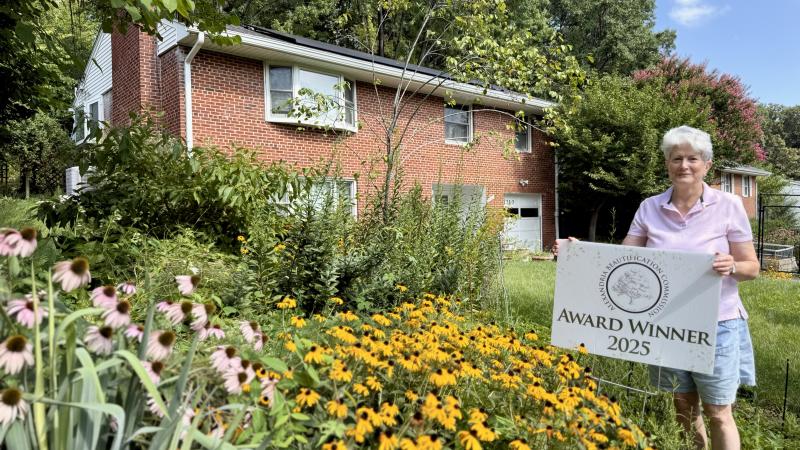 Woman smiling behind a yard sign that says Award Winner 2025 in front of a brick house surrounded by colorful flowers and green shrubs and trees