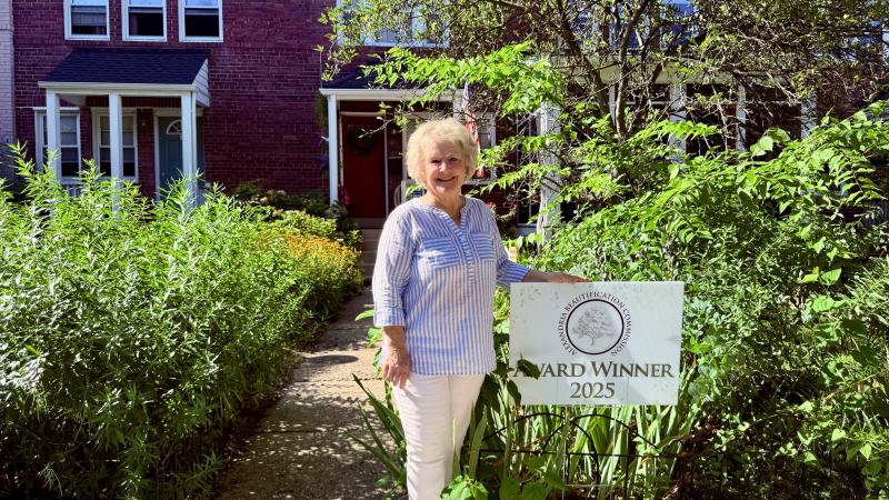 Woman stands next to a sign that says Award Winner 2025 in front of her yard full of green shrubbery and trees