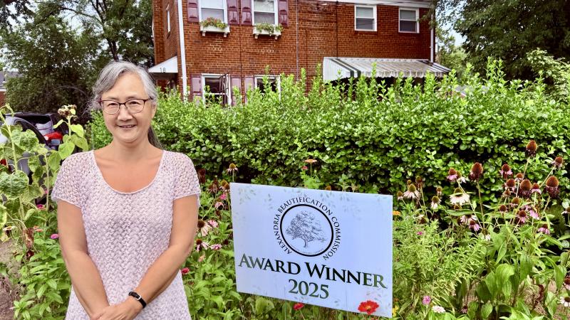 Image a woman standing next to a sign that says Award Winner 2025 surrounded by greenery and a brick house in the back