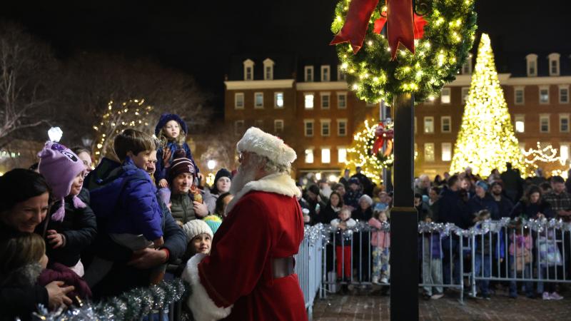 A man dressed as Santa greets a young child. A lighted holiday tree and City Hall can be seen in the background.