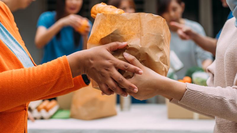 A woman hands a bag of donated food to someone during a community food drive. A female volunteer and her colleagues distributing grocery food at community food bank