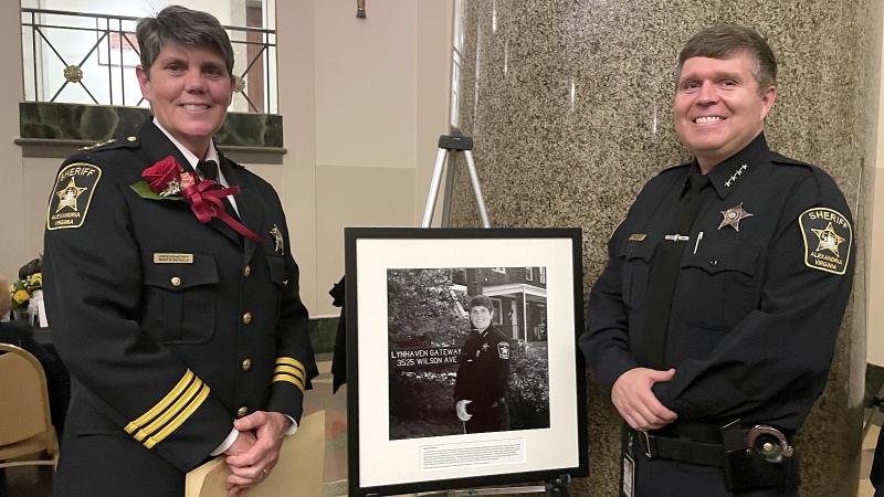 Undersheriff on left and Sheriff on right, both in blue uniforms, standing with a framed black and white photograph of the undersheriff between them