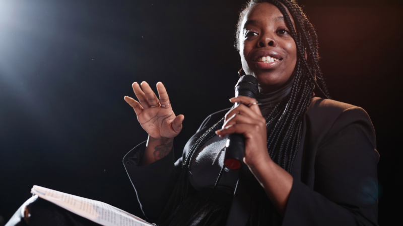 Image of a black woman holding a mic on a black background and a spotlight coming from the top left