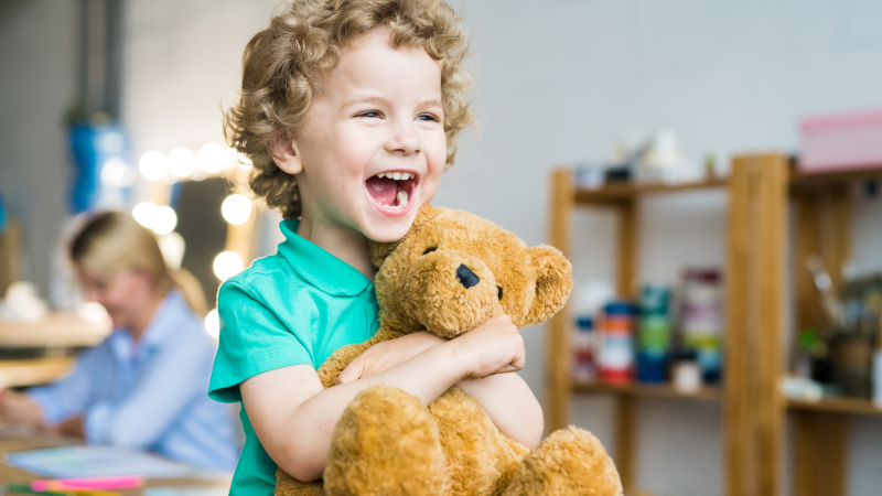 Image of a curly haired dirty blonde boy smiling with mouth wide open wearing a green shirt while hugging a brown teddy bear set in an indoor recreation facility that's blurred in the background