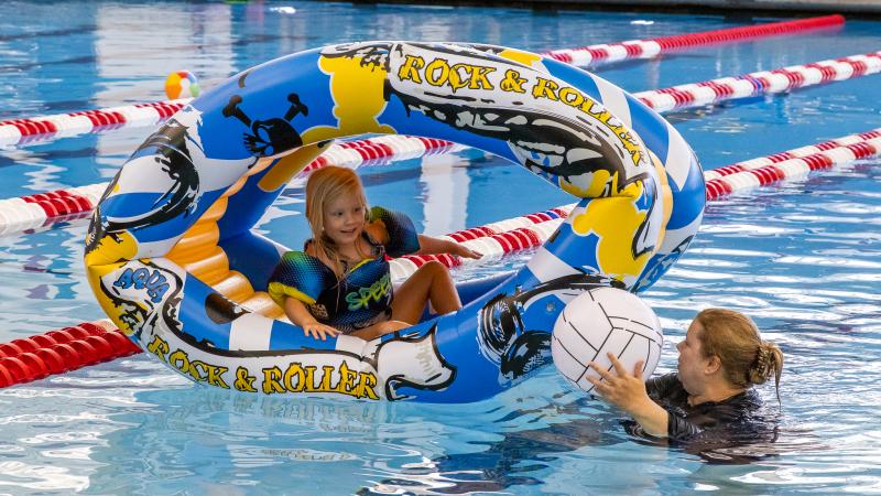 A child sitting inside one blow up pool toy while her mother, who's standing next to her, hands her a blow up volleyball pool toy