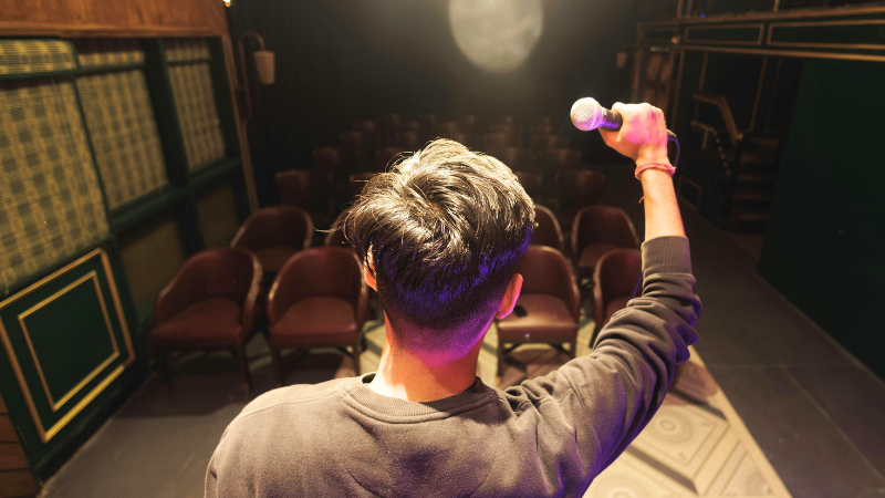A man holds a microphone facing a large, empty room 