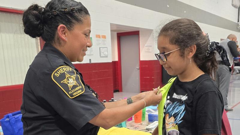 Sheriff's sergeant in blue uniform pinning a badge on the safety patrol belt of an elementary school student at a community event
