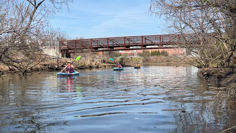 Three kayakers one after the other on the river with paddles, just went under a bridge at Four Mile Run Park