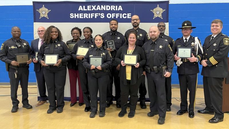 Deputies and civilian staff holding plaques and smiling at an awards ceremony in front a backdrop that says Alexandria Sheriff's Office