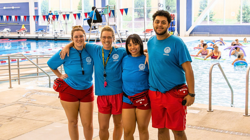 Four lifeguards wearing blue shirts and red shorts with arms around each other and smiling for the camera at Minnie Howard Aquatics Facility