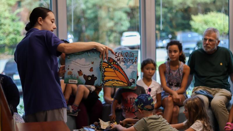 Nature Center staff member is holding a book open while standing and showing it to community members who are sitting and listening to the story time