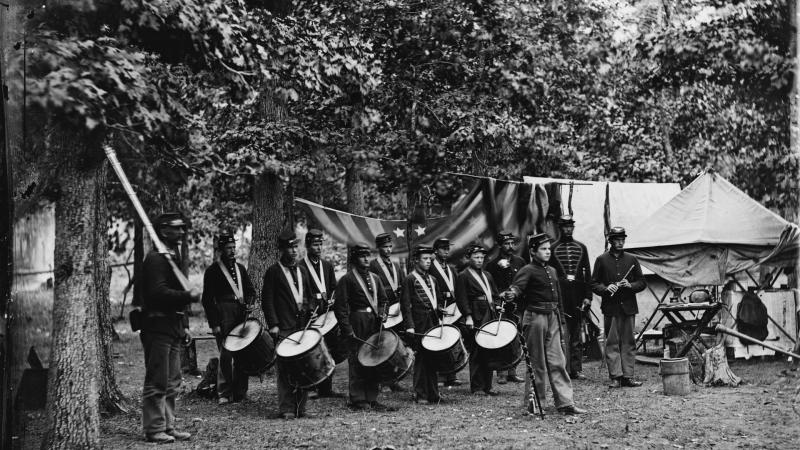 The drum corps poses with their drums, in camp beside a draped American flag and tents.