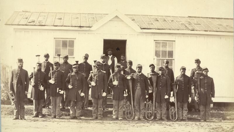 African American soldiers pose with their instruments in front of a long white building.