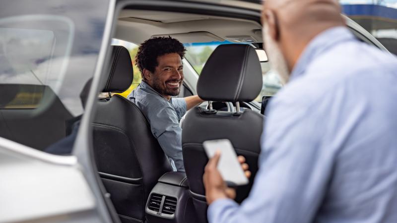 A man holds a cell phone and looks inside the back seat of a car. The driver looks back and smiles. 