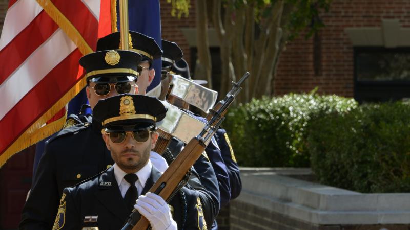 A public safety honor guard in formal uniforms carry ceremonial rifles and an American flag in a procession.